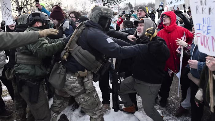 Protestors and armed federal agents struggle in close quarters, with shouting and signs visible amid a snowy street demonstration.