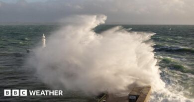 A large wave crashes against a breakwater with a lighthouse sending up a big spray of foam