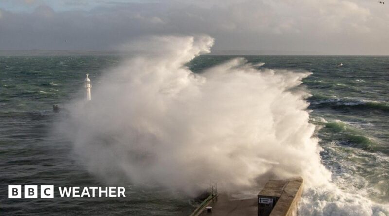 A large wave crashes against a breakwater with a lighthouse sending up a big spray of foam