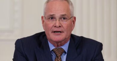 Darren Woods speaks at a meeting, wearing a dark suit, light blue shirt and patterned brown tie.