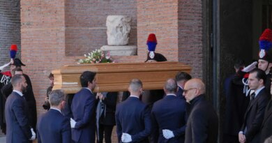 A wooden casket with white and pink flowers is carried by people in dark suits. A brick wall and uniformed people with red and blue hats are in the background.