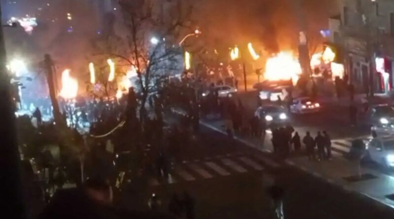 A blurry image of a dark street filled with fires, cars and people.