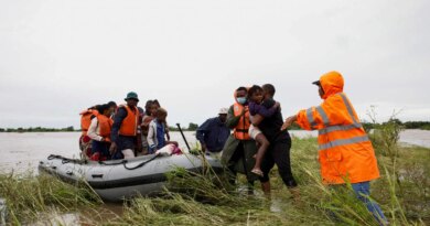 A few adults, one carrying a child, wade through flooded vegetation toward an inflatable boat that already has several people in it.