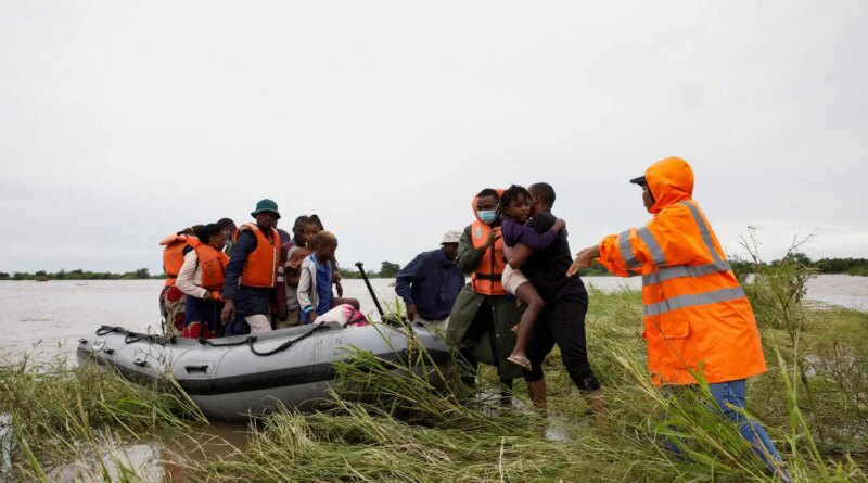 A few adults, one carrying a child, wade through flooded vegetation toward an inflatable boat that already has several people in it.