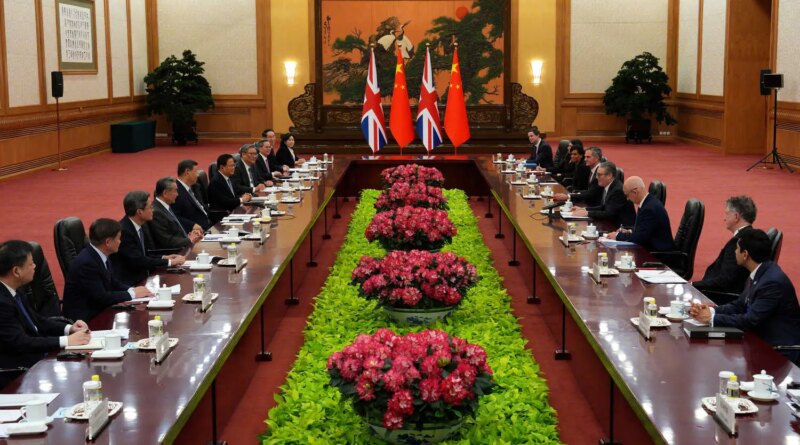A formal meeting with two rows of delegates, all in dark suits, facing each other across a long table. There are Chinese and British flags in the back.
