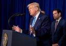 Donald Trump speaks at a podium with the presidential seal, gesturing with both hands, as another man stands behind him.