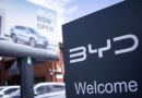 A BYD showroom sign in the foreground with a billboard showing a BYD car and the words "Now open" in the background.