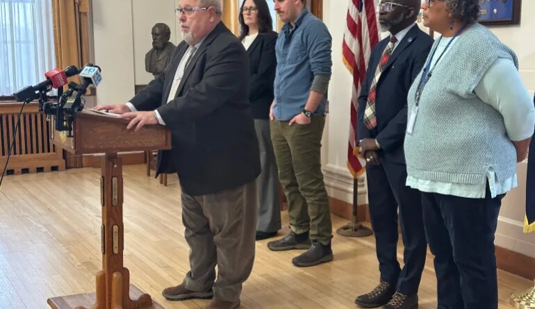 Mark Dion speaks at a podium, surrounded by city officials