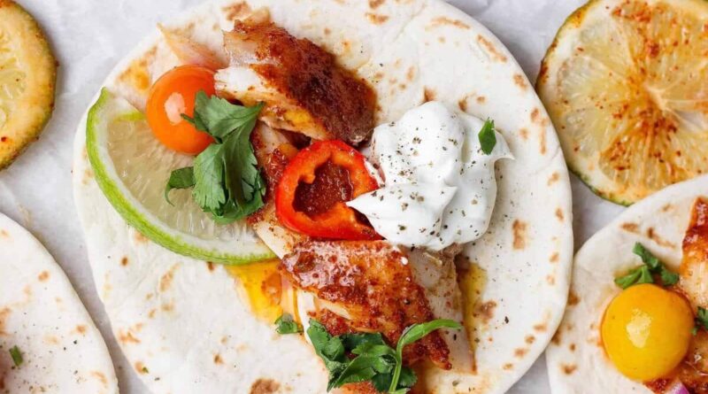 A soft flour tortilla topped with grilled fish, sliced lime, cherry tomato, red pepper, fresh parsley, and a dollop of sour cream. Grilled lime slices are visible nearby.