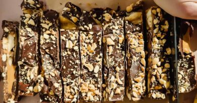 Sliced chocolate bars topped with chopped nuts are arranged on parchment paper, with a hand holding a knife to cut one of the bars.