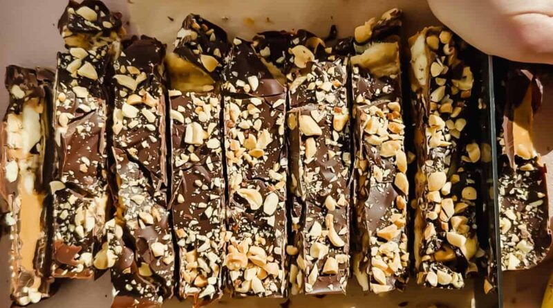 Sliced chocolate bars topped with chopped nuts are arranged on parchment paper, with a hand holding a knife to cut one of the bars.
