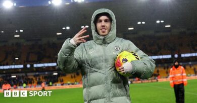 Cole Palmer celebrates scoring a hat-trick for Chelsea with a three-finger salute with his right hand, while holding the yellow match ball under his left arm and wearing a large Chelsea puffer jacket with the hood up.