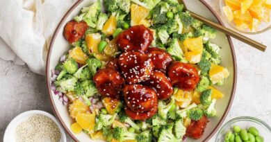 A bowl filled with broccoli, orange pieces, edamame, red onion, and glazed meatballs topped with sesame seeds, with small bowls of orange slices, edamame, and sesame seeds nearby.