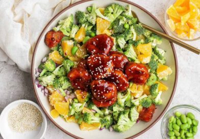 A bowl filled with broccoli, orange pieces, edamame, red onion, and glazed meatballs topped with sesame seeds, with small bowls of orange slices, edamame, and sesame seeds nearby.
