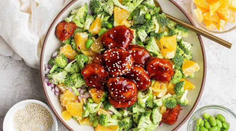 A bowl filled with broccoli, orange pieces, edamame, red onion, and glazed meatballs topped with sesame seeds, with small bowls of orange slices, edamame, and sesame seeds nearby.