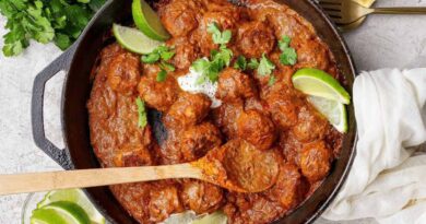 A cast iron skillet filled with butter chicken meatballs in a rich red sauce, garnished with lime wedges and cilantro, with a wooden spoon resting inside. Fresh cilantro and lime are on the side.