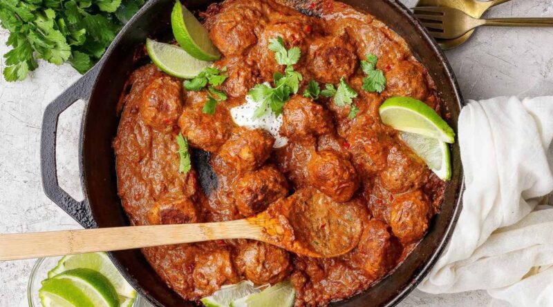 A cast iron skillet filled with butter chicken meatballs in a rich red sauce, garnished with lime wedges and cilantro, with a wooden spoon resting inside. Fresh cilantro and lime are on the side.