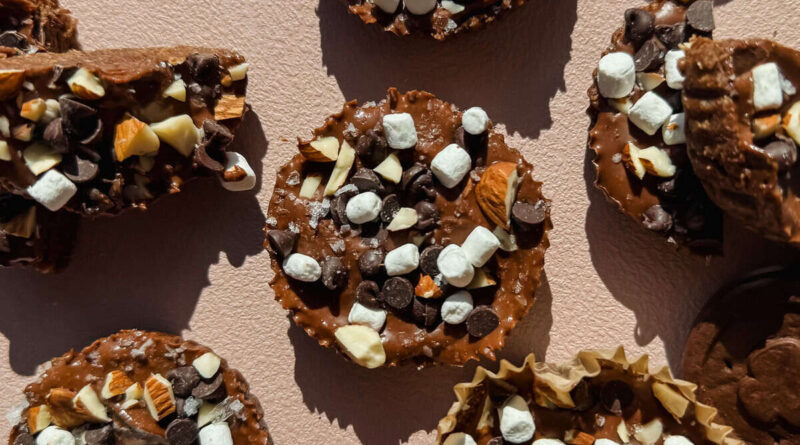 Chocolate treats topped with mini marshmallows, chocolate chips, and chopped nuts are arranged on a light textured surface.