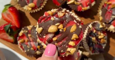 A hand with pink nail polish holds a chocolate-covered treat topped with chopped nuts and strawberry slices, with more treats and strawberries visible on a wooden tray.