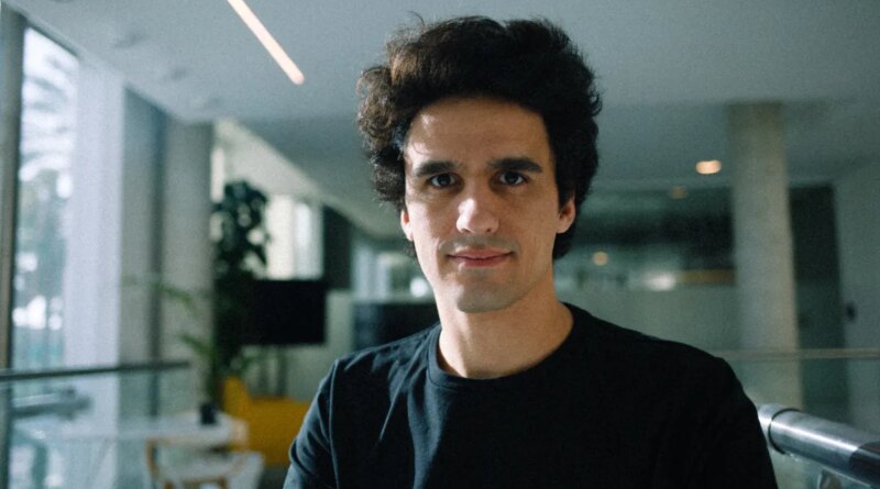 A man wearing a large black t-shirt stands in a sunlit office.