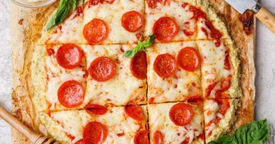 A round chicken crust pizza with melted cheese and pepperoni, cut into square slices and garnished with basil, sits next to a knife and a small bowl of grated cheese.