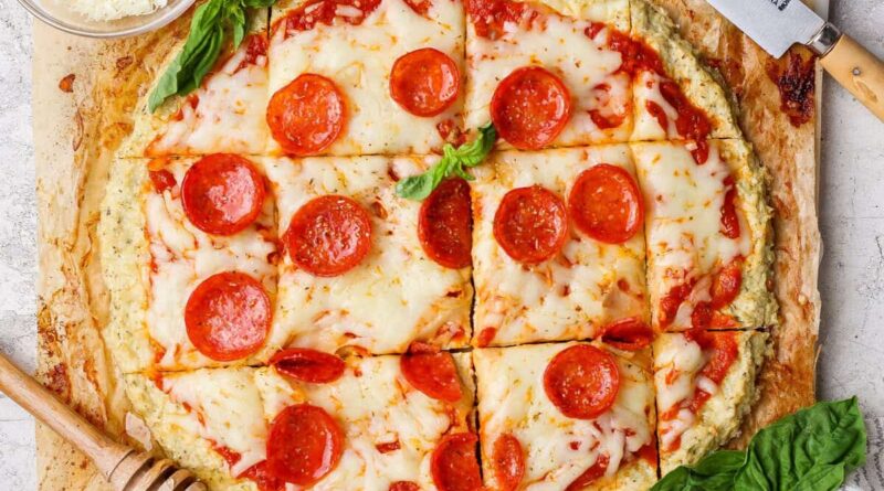 A round chicken crust pizza with melted cheese and pepperoni, cut into square slices and garnished with basil, sits next to a knife and a small bowl of grated cheese.