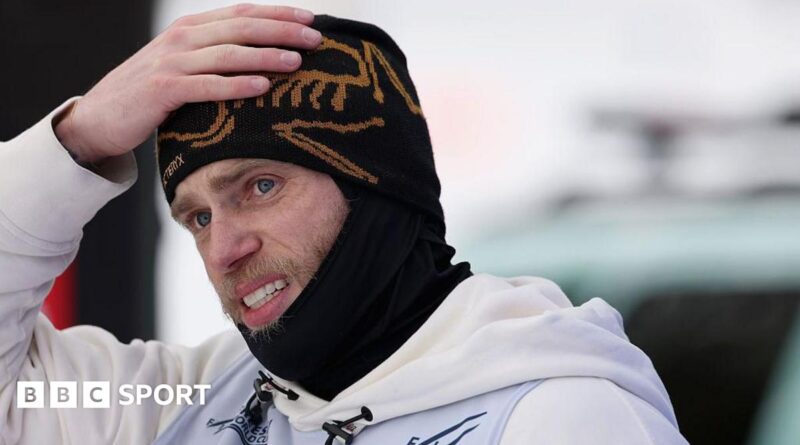 Gus Kenworthy is wearing a black beanie and snood and has his hand on his head