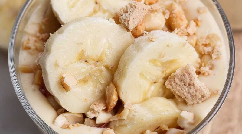 A glass bowl filled with banana pudding, topped with banana slices, chopped nuts, and crumbled cookies.
