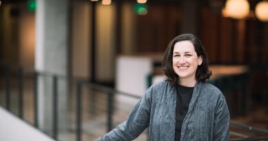 A dark-haired woman poses by a balcony in a gray cardigan sweater.
