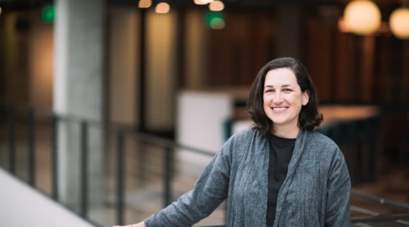 A dark-haired woman poses by a balcony in a gray cardigan sweater.