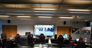 A gaming room at the University of Sussex, featuring multiple gaming stations with black and red chairs, a central table with green pamphlets, and a large screen displaying a video game.
