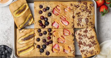 A sheet pan divided into six baked pancake squares, each topped with different ingredients like banana, blueberries, strawberries, peanut butter, and chocolate pieces.