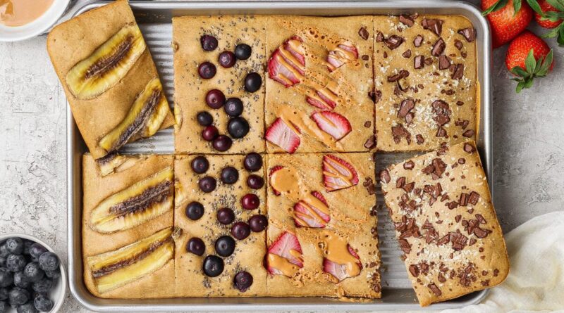 A sheet pan divided into six baked pancake squares, each topped with different ingredients like banana, blueberries, strawberries, peanut butter, and chocolate pieces.