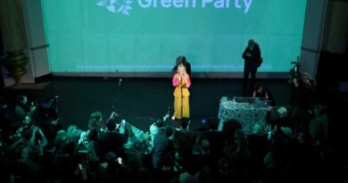 A woman dressed in yellow and pink speaks underneath a large screen with a Green Party emblem projected on it.