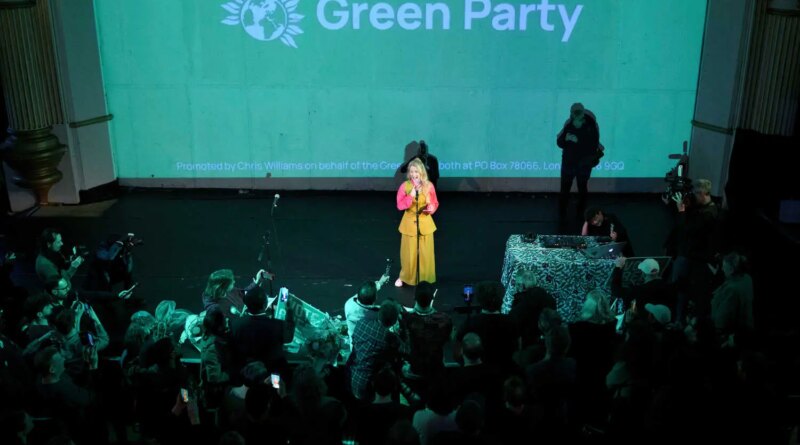 A woman dressed in yellow and pink speaks underneath a large screen with a Green Party emblem projected on it.