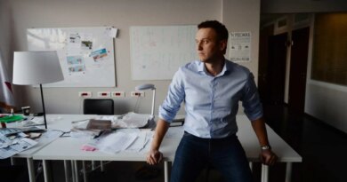 Aleksei A. Navalny sits on a white desk in a cluttered office. He wears a light blue shirt and dark pants, looking away with a neutral expression.