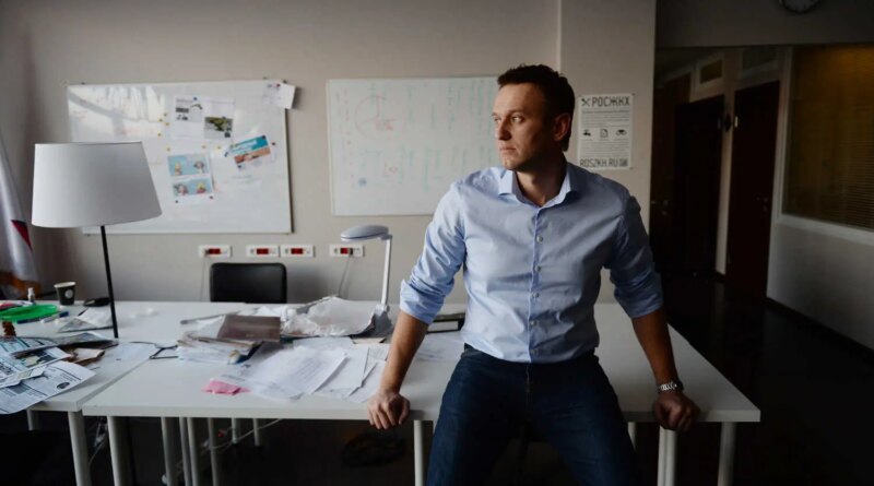 Aleksei A. Navalny sits on a white desk in a cluttered office. He wears a light blue shirt and dark pants, looking away with a neutral expression.