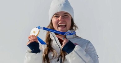 Breezy Johnson, wearing a white beanie hat and a white coat, smiling and holding up her gold medal.