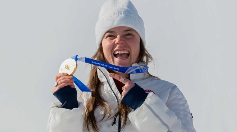 Breezy Johnson, wearing a white beanie hat and a white coat, smiling and holding up her gold medal.