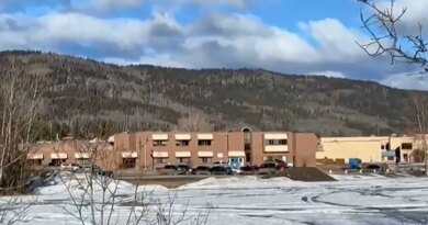 A school building with cars and snow out front. There are hills in the background.