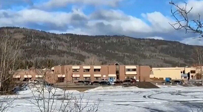 A school building with cars and snow out front. There are hills in the background.