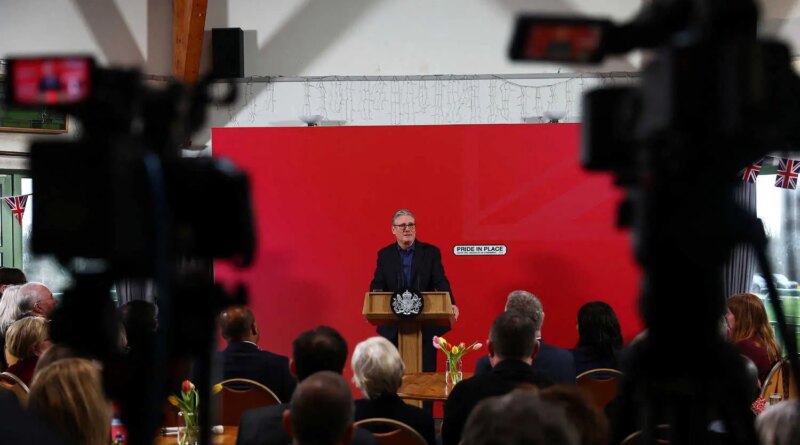 Keir Starmer speaks at a lectern, facing an audience. Behind him is a red wall with "PRIDE IN PLACE" and Union Jack flags.