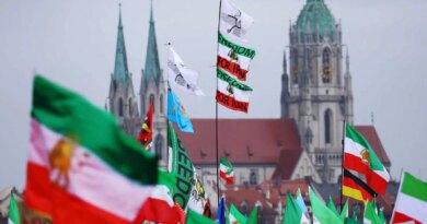 Numerous green, white and red flags, some reading "FREEDOM FOR IRAN," waving in front of two old stone church spires, on the left, and a domed building.