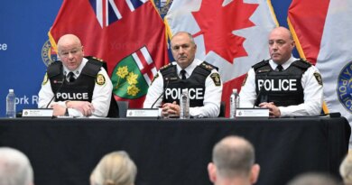 Three police officers sit at a table in front of microphones. Flags hang behind them.