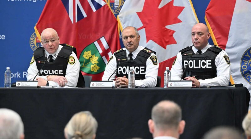 Three police officers sit at a table in front of microphones. Flags hang behind them.
