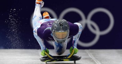 A man holding a skeleton sled on an ice track, wearing a helmet with images of people on it.