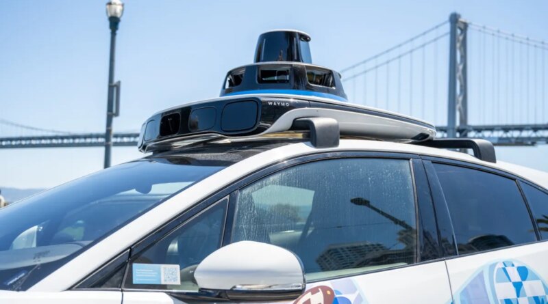Self-driving Waymo car with rooftop lidar and bird-themed mural on the Embarcadero with the San Francisco-Oakland Bay Bridge in background, San Francisco, California, August 14, 2025.