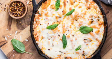 A cast iron skillet filled with baked cheesy pasta topped with melted cheese, fresh basil leaves, and sprinkled red pepper flakes. The skillet is on a wooden board with a small bowl of red pepper flakes and basil nearby.
