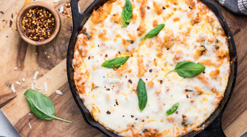 A cast iron skillet filled with baked cheesy pasta topped with melted cheese, fresh basil leaves, and sprinkled red pepper flakes. The skillet is on a wooden board with a small bowl of red pepper flakes and basil nearby.