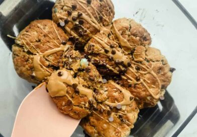 A glass dish containing seven chunky cookies drizzled with light brown icing and sprinkled with mini chocolate chips, one cookie being lifted with a pink spatula.